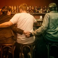Foto von drei Menschen an einer Bar. Alle drei sitzen auf Barhockern, wir sehen nur ihre Rücken. Sie umarmen sich alle drei miteinander.