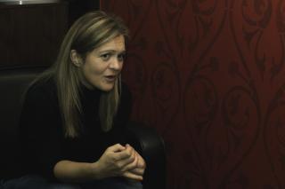 Foto von Barbara Eder in einem schwarzen Sessel vor einer roten, gemusterten Wand. Barbara Eder schaut konzentriert, Interviewsituation.